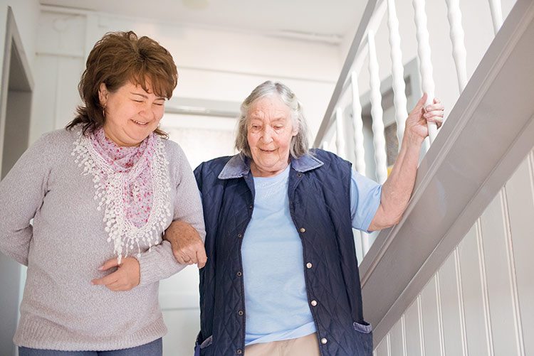 Eine ältere Dame steht mit ihrer Pflegerin lächelnd in einem Treppenhaus