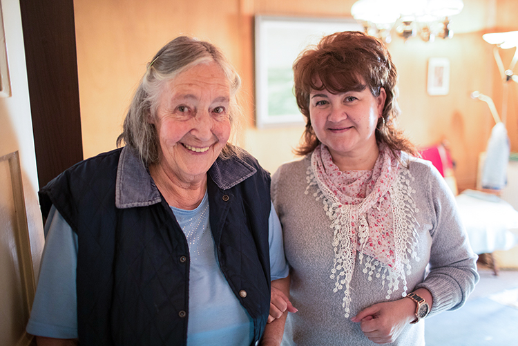 Eine ältere Dame und ihre Pflegerin stehen in einer Wohnung und lächeln in die Kamera.